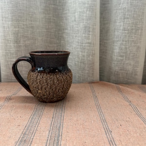 May include: A handcrafted ceramic mug with a dark brown glazed rim and handle. The lower part of the mug has a textured, earthy brown finish. The mug is placed on a peach-coloured fabric with blue stripes, against a neutral backdrop.