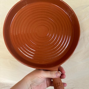 May include: A terracotta-coloured, round ceramic pan with a handle. The pan has a wide, shallow bowl with concentric circular ridges. The handle is integrated and features a decorative design at the end. The pan is held by a person.