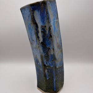 May include: A tall, blue ceramic vase with an unusual, angular form. The vase displays a mix of blue shades with darker, textured sections, producing a marbled appearance. The base is a lighter, natural tone, and the vase has a gently curved design.