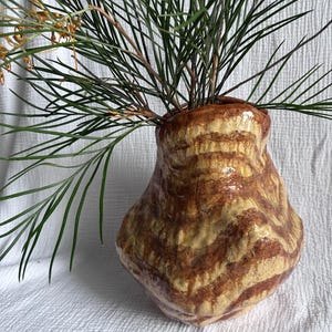 May include: A handmade ceramic vase with a unique, organic shape. The vase features a brown and tan striped pattern and holds green and yellow plant stems. The vase sits on a white textured surface.