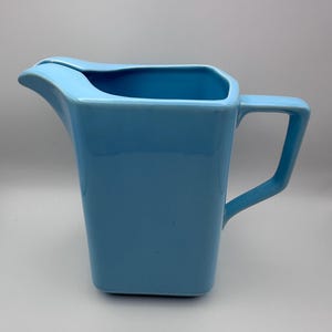 May include: A light blue ceramic jug with a rectangular body and a curved spout. The jug has a handle and a capacity of approximately 1.9 liters. The design is simple and modern, suitable for serving beverages.