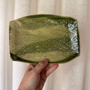 May include: A rectangular ceramic serving dish with a raised floral pattern in shades of green. The dish is held by a hand, showcasing its decorative design and shape. The edges are slightly rounded, and the overall aesthetic is rustic.