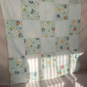 May include: A square patchwork quilt featuring an alternating grid pattern of solid light blue fabric squares and floral print squares. The floral fabric displays a dense pattern of small yellow, red, and blue flowers with green leaves on a white background.