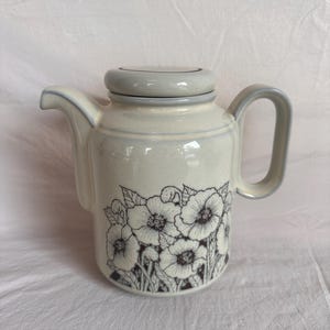 May include: Cream ceramic teapot with a grey lid and handle. The teapot has a black and white floral design on the front. The spout and handle are outlined in grey.