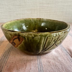 May include: A green ceramic bowl with a leaf design. The bowl has a glossy finish and a unique, aged appearance. A small, brown and tan design is on the side of the bowl. The bowl is on a pink and white striped surface.