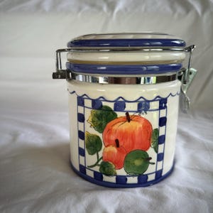 May include: A ceramic storage jar with a lid and metal clasp. The jar is white with a blue chequered pattern and a painted design of pumpkins and green leaves. The lid has blue and white stripes.