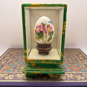 May include: A white egg-shaped ornament featuring hand-painted pink flowers and a small blue bird, resting on a dark brown wooden stand. The ornament is enclosed within a rectangular green fabric-covered display case with gold floral patterns.