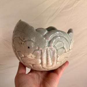 May include: A ceramic bowl with a distinctive, textured pattern. The bowl is mainly a pale green, with a lighter, almost white, base. The rim has a scalloped edge, enhancing its decorative look. The bowl is held in a hand.