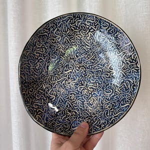 May include: A circular ceramic plate featuring a dark blue base with a white, abstract design. The plate has a black rim and is held by a hand. The plate is likely used for serving food, and is set against a white curtain.
