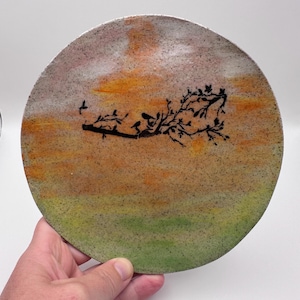 May include: Circular decorative plate featuring a painted sunset in orange, yellow, and green hues. A black silhouette of a tree branch with birds is painted across the centre. The plate is held by a hand.