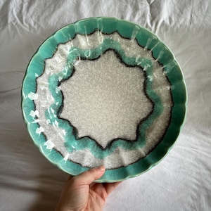 May include: A ceramic plate with a scalloped edge. The plate has a turquoise rim, a white and grey speckled centre, and a dark brown accent line. The plate is held by a hand.