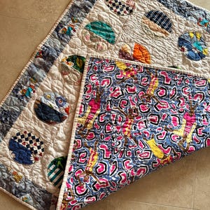 May include: A handmade quilt with a cream-coloured top featuring appliquéd circles in various patterns and colours, including blue, green, and orange. The quilt's border is a blue and white pattern. The underside reveals a vibrant, patterned fabric.