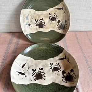 May include: Two ceramic plates with a Japanese-inspired design. Each plate features a dark green rim, a beige and white central band, and a crab motif. The plates are round and suitable for serving food.