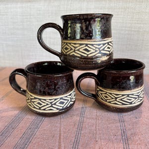 May include: Three dark brown ceramic mugs with a cream geometric pattern. The mugs have rounded handles and are stacked. The mugs are on a pink and white striped surface. The mugs are suitable for tea or coffee.