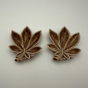May include: Two brown ceramic leaf-shaped ornaments. Each leaf features detailed veining and a glossy finish. The leaves are displayed on a white surface, highlighting their intricate design and earthy tones. These pieces are likely for home decor.