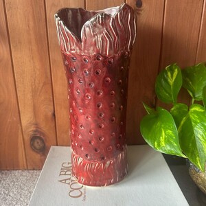 May include: A tall, red ceramic vase with a textured surface and a unique, scalloped top. The vase features small, circular indentations and white trim. The vase is displayed on a stack of books.