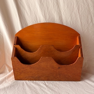 May include: A wall-mounted wooden mail organiser featuring a warm, medium-brown wood grain finish. The unit has two tiered compartments with scalloped front edges and a rounded, arched back panel for mounting, providing storage for letters.