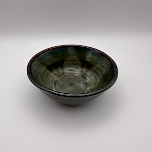 May include: A handcrafted ceramic bowl with a dark green and brown glaze. The bowl has a rounded form with a slightly flared rim. The interior features a swirling pattern of green and brown hues, creating a unique aesthetic. Suitable for serving food or as a decorative piece.