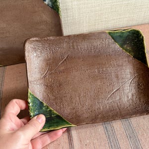 May include: Rectangular brown ceramic plates with textured surfaces and green glazed corners. The plates have a rustic, handmade appearance, with subtle etched designs. The plates are suitable for serving food.