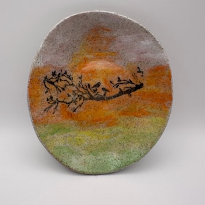 May include: Handmade ceramic plate with a painted landscape design. The plate features a black tree branch with birds against a backdrop of orange, yellow, and green hues, with a speckled grey rim.