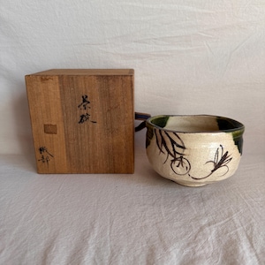 May include: A ceramic tea bowl with a cream-coloured base, featuring dark green glaze accents on the rim and black hand-painted floral motifs on the exterior. The bowl is accompanied by a square wooden storage box with black Japanese calligraphy.
