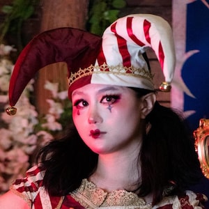 May include: A jester's hat with a burgundy and white color scheme, featuring red stripes and gold accents. The hat has a bell at the end of each point. The person in the image is wearing makeup with a star design.
