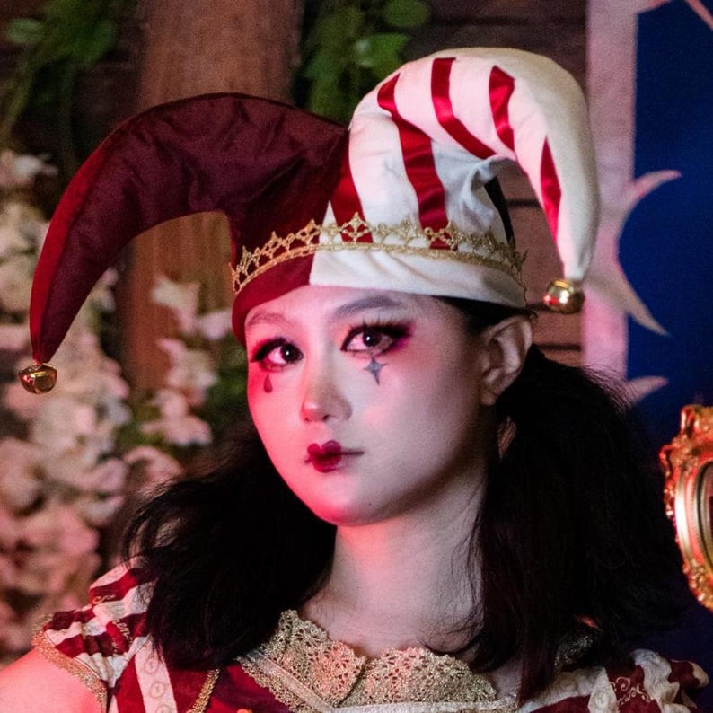 May include: A jester's hat with a burgundy and white color scheme, featuring red stripes and gold accents. The hat has a bell at the end of each point. The person in the image is wearing makeup with a star design.