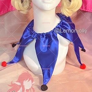May include: A royal blue satin jester collar featuring a gathered neckline and multiple pointed panels. Each point of the collar is finished with a small, round, red pom-pom. The collar is displayed on a white mannequin bust with blonde hair.