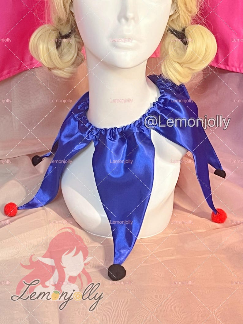 May include: A royal blue satin jester collar featuring a gathered neckline and multiple pointed panels. Each point of the collar is finished with a small, round, red pom-pom. The collar is displayed on a white mannequin bust with blonde hair.