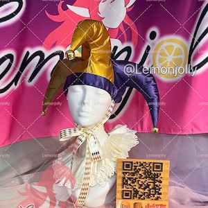 May include: A jester's hat with gold, brown, and purple panels, adorned with small bells. The hat is displayed on a mannequin head, paired with a white ruffled collar and a checkered ribbon. A wooden block with a QR code and social media handles is in the foreground.