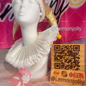 May include: A white pleated ruff collar with delicate lace trim worn by a white mannequin head. The mannequin wears a gold jester hat with small yellow bells. A wooden sign with a QR code and the text Etsy, Instagram, and Lemonjolly is present.