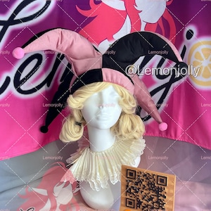 May include: A jester costume set featuring a pink and black harlequin hat with pink pom-poms, a ruffled collar, and a blonde wig on a mannequin head. A wooden sign with a QR code and social media handles is in front.