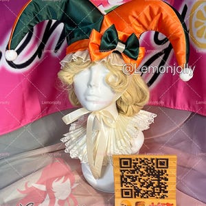 May include: A jester hat with green and orange fabric, white pom-poms, and a satin bow. The hat is displayed on a mannequin head with a blonde wig and a ruffled collar. A wooden sign with a QR code and social media handles is in front.