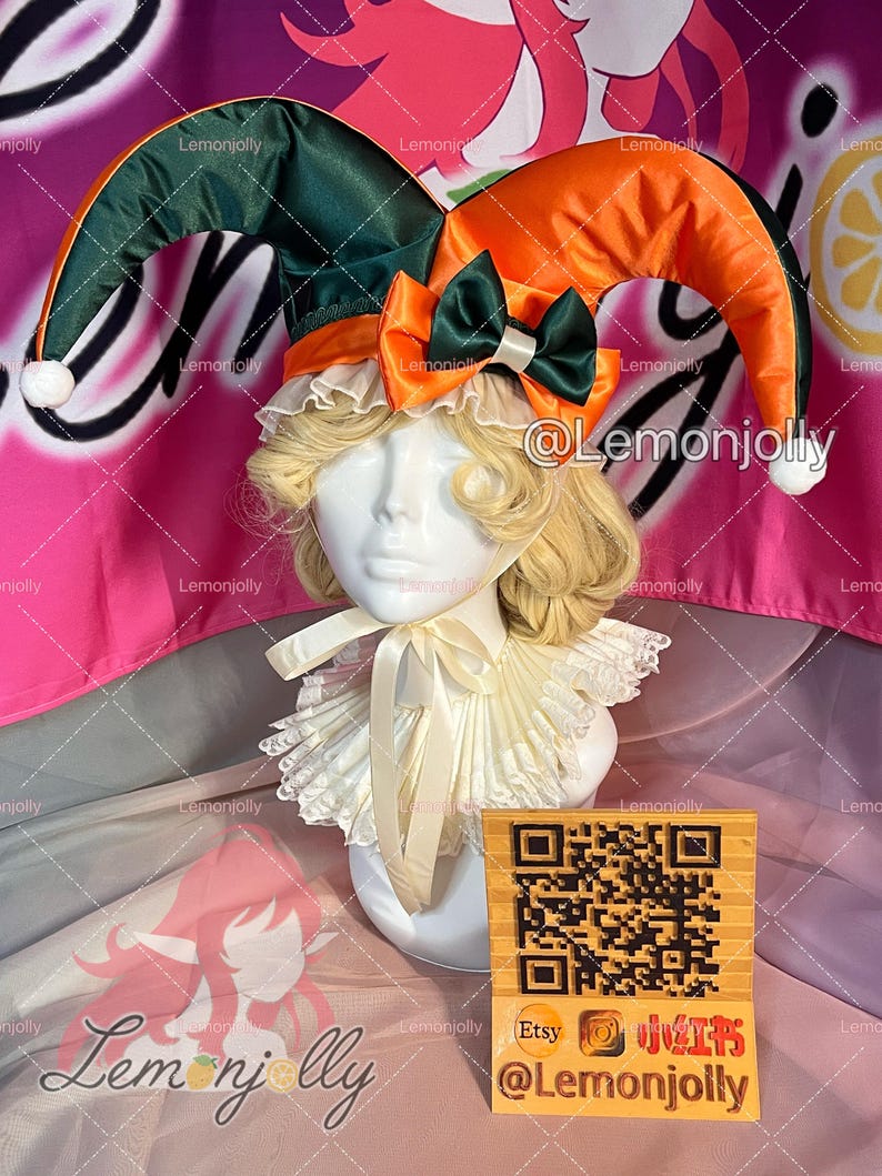 May include: A jester hat with green and orange fabric, white pom-poms, and a satin bow. The hat is displayed on a mannequin head with a blonde wig and a ruffled collar. A wooden sign with a QR code and social media handles is in front.