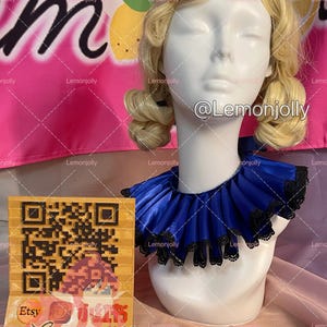 May include: A white mannequin head with blonde hair wearing a royal blue satin ruff collar featuring a black lace trim along the edges. The collar is pleated and sits around the neck of the mannequin. A square card with a QR code is visible.