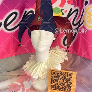 May include: A jester's hat with burgundy and navy blue panels, adorned with small bells. A white ruffled collar and a wooden sign with a QR code and social media handles are also visible. The background is pink and white.