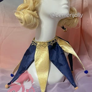 May include: A jester collar featuring alternating navy blue velvet and gold satin points. The navy blue points have a subtle star pattern. The collar is accented with a gold lace trim at the neck and small blue and beige pom-poms at each tip. Text reads @Lemonjolly.