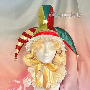 May include: A mannequin head wearing a jester's hat and ruffled collar. The hat is red, green, and gold with bells at the tips. The collar is cream-colored with a gold ribbon. The wig is blonde with curly locks.