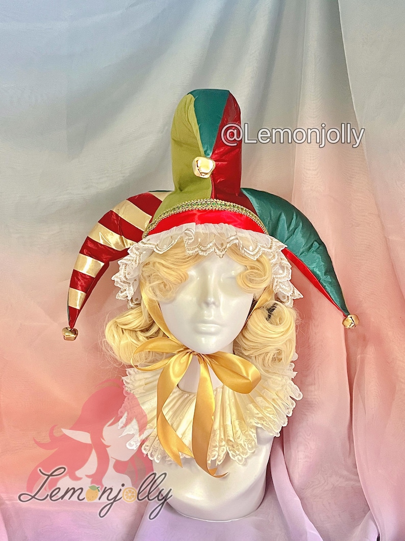 May include: A mannequin head wearing a jester's hat and ruffled collar. The hat is red, green, and gold with bells at the tips. The collar is cream-colored with a gold ribbon. The wig is blonde with curly locks.