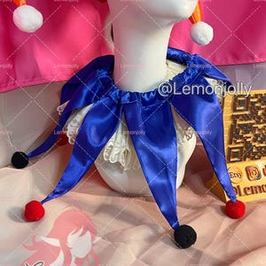 May include: A jester collar made of royal blue satin fabric with pointed ends, each tipped with a small red or black pom-pom. The collar is layered over a white lace ruff and is displayed on a white mannequin head wearing a red and orange hat.