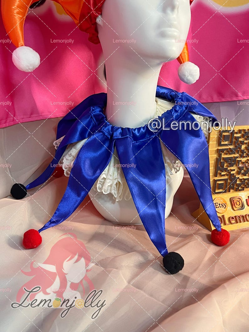 May include: A jester collar made of royal blue satin fabric with pointed ends, each tipped with a small red or black pom-pom. The collar is layered over a white lace ruff and is displayed on a white mannequin head wearing a red and orange hat.