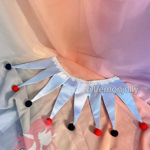 May include: A light blue jester collar featuring a white lace trim along the neckline and alternating black and red pom-poms at the tips of each pointed segment. The text @Lemonjolly is visible on the right side of the collar.
