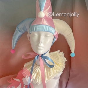 May include: A jester's hat and collar set. The hat is pink and blue with white pom-poms and long, pointed tips. The collar is cream-colored with a ruffled design and a pink and blue ribbon. The text "@Lemonjolly" is visible.