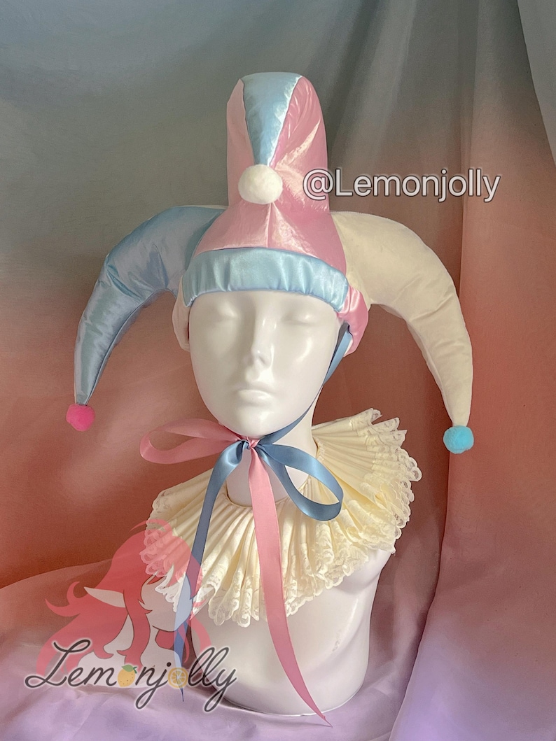 May include: A jester's hat and collar set. The hat is pink and blue with white pom-poms and long, pointed tips. The collar is cream-colored with a ruffled design and a pink and blue ribbon. The text "@Lemonjolly" is visible.