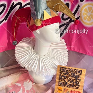 May include: A jester's hat with red, gold, yellow, and teal accents, atop a white mannequin head. The hat has a red velvet band and dangling pom-poms. A white ruffled collar is around the neck. A wooden sign with a QR code is also present.