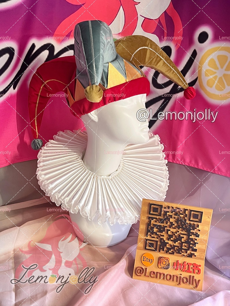 May include: A jester's hat with red, gold, yellow, and teal accents, atop a white mannequin head. The hat has a red velvet band and dangling pom-poms. A white ruffled collar is around the neck. A wooden sign with a QR code is also present.
