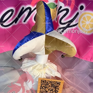 May include: A multi-colored jester hat with a white ruffled collar. The hat features sections of blue, gold, and white fabric. A small wooden sign with a QR code and text is in front of the collar. The background is pink with text.