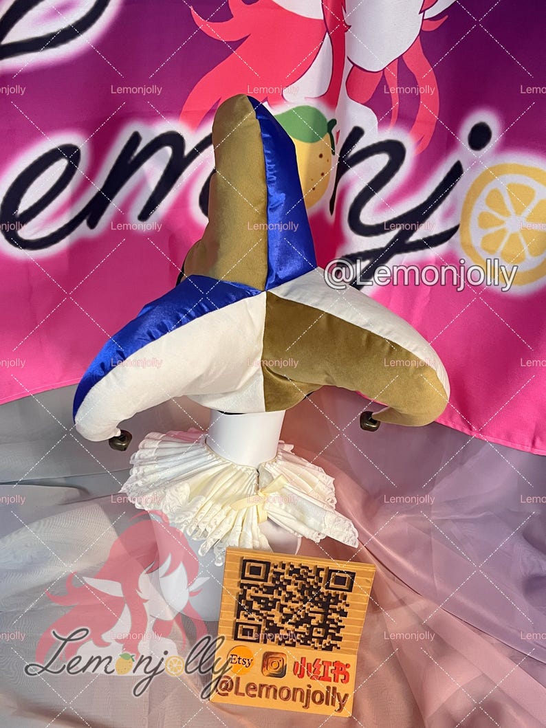 May include: A multi-colored jester hat with a white ruffled collar. The hat features sections of blue, gold, and white fabric. A small wooden sign with a QR code and text is in front of the collar. The background is pink with text.