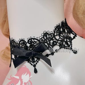 May include: A black lace choker necklace featuring an intricate floral pattern with scalloped edges and a small black satin ribbon bow attached to the front. The design includes delicate dangling lace accents and the text @Lemonjolly.