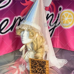May include: A tall, conical headpiece in light blue and cream, featuring a delicate white lace trim, a string of pearls along the seam, and a sheer, flowing veil. The headpiece is topped with a small gold bell and includes cream-colored ribbons.